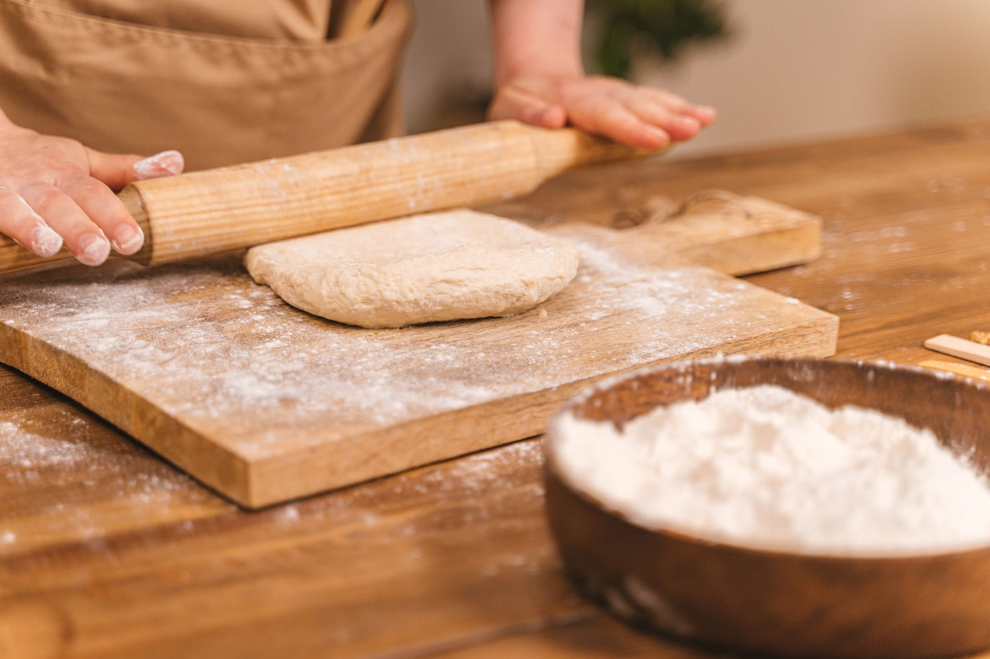 Close-up of hands kneading dough with a rolling pin on a floured wooden board for home baking.