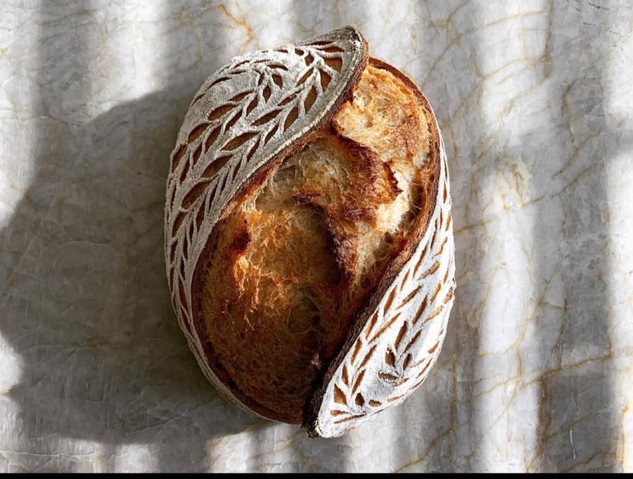 A round loaf with an intricate decorative leaf pattern scored across half of its crust, cut open to show the crumb
