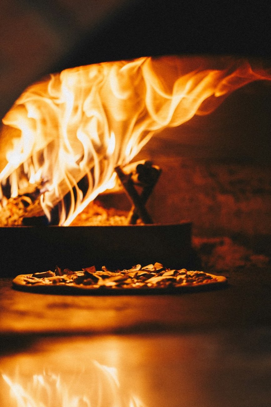 Flames inside a wood-fired pizza oven with a pizza baking on the hot stone floor, illustrating the home oven heat chapter